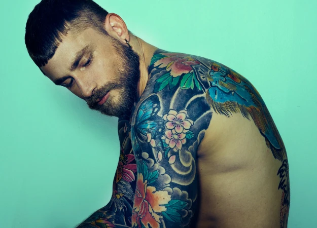 Adult male with beard, short hair, partially shirtless with body tattoos, posing indoors against a light blue background in studio lighting.