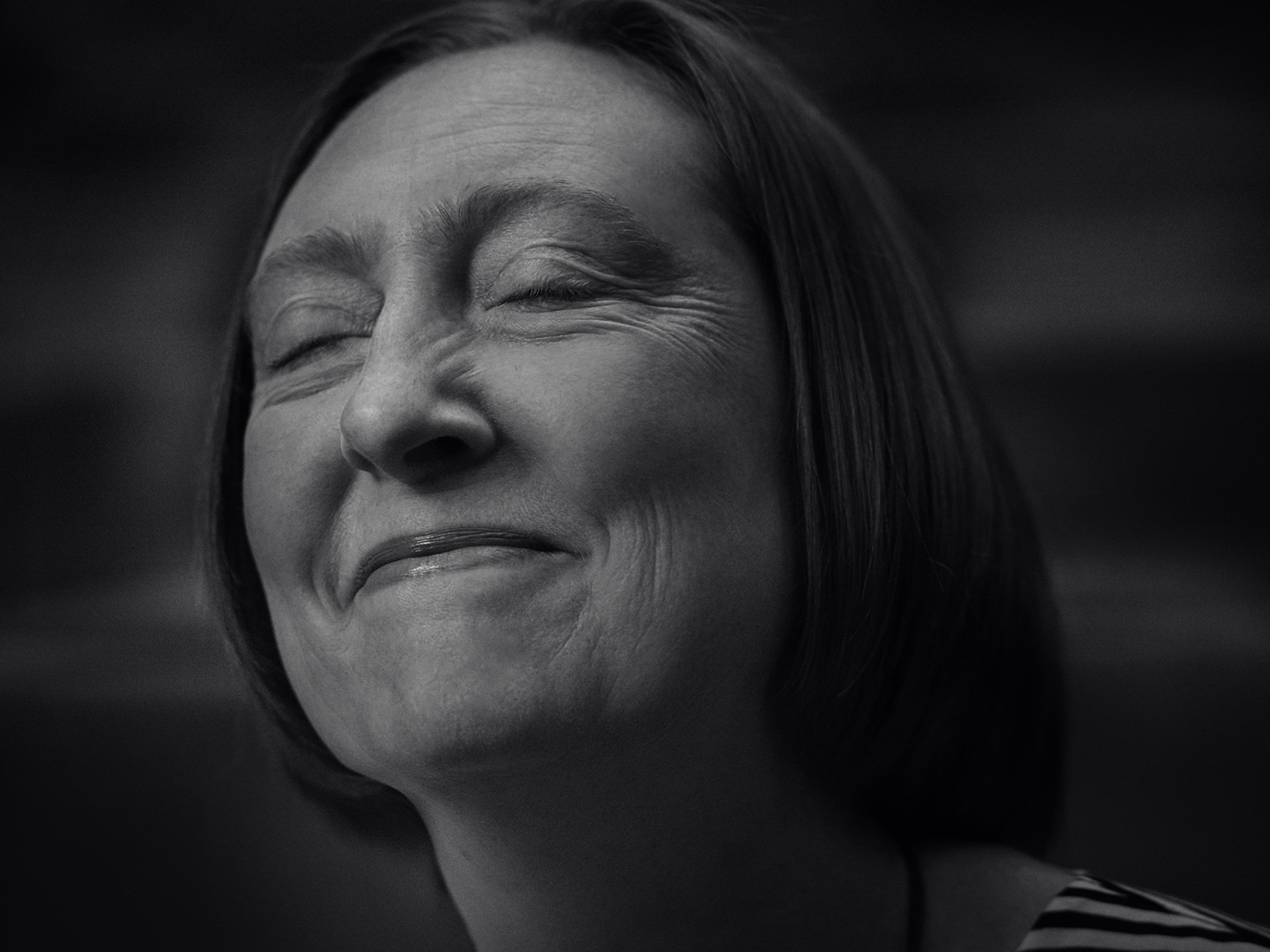 Monochrome close-up portrait of a mature woman with closed eyes, wearing a striped shirt, against a dark backdrop.