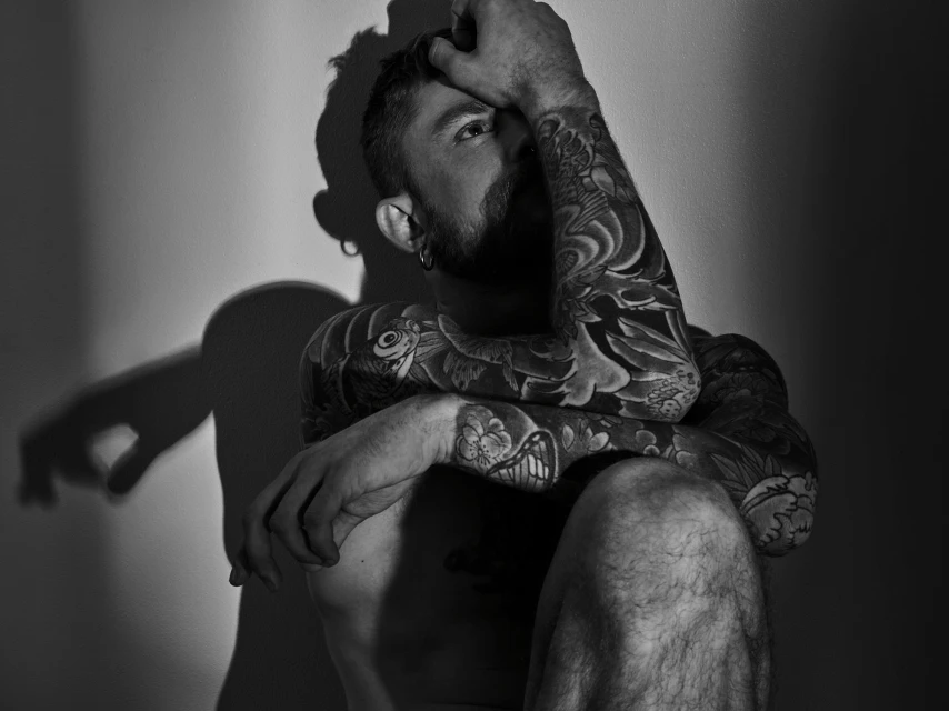 Monochrome portrait of a tattooed adult male posing with hands on his head in front of a wall in studio lighting.