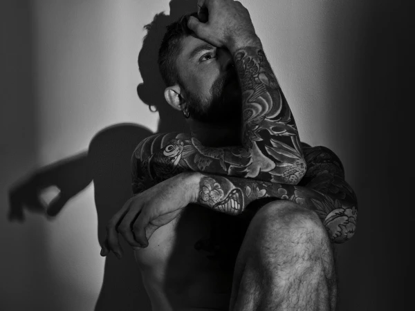 Monochrome portrait of a tattooed adult male posing with hands on his head in front of a wall in studio lighting.