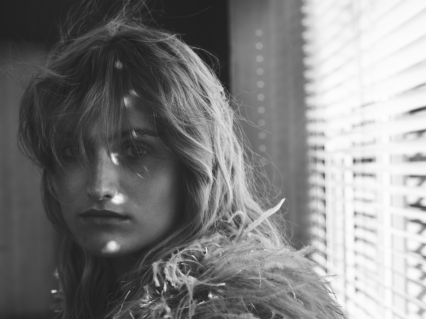 Black and white portrait of a woman with feathered clothing standing by blinds, creating shadows on her face in an indoor environment.