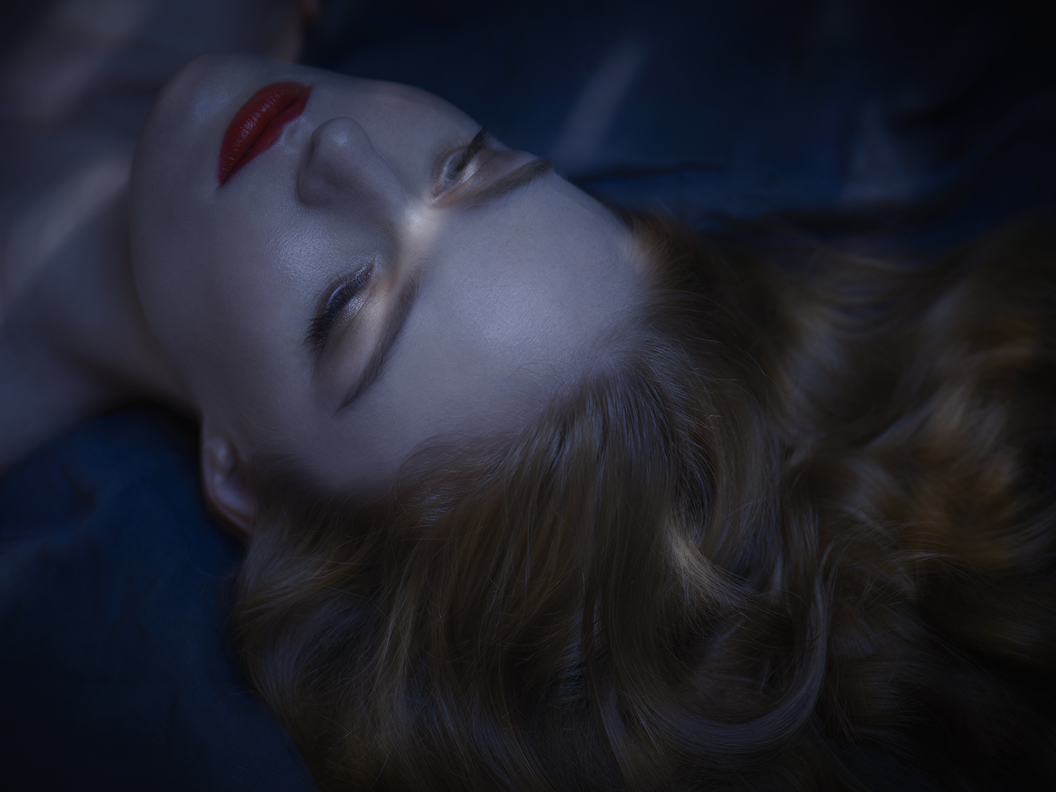 Close-up of a woman with fair skin and red lipstick lying down with reddish-brown wavy hair, illuminated with a blue and white light.