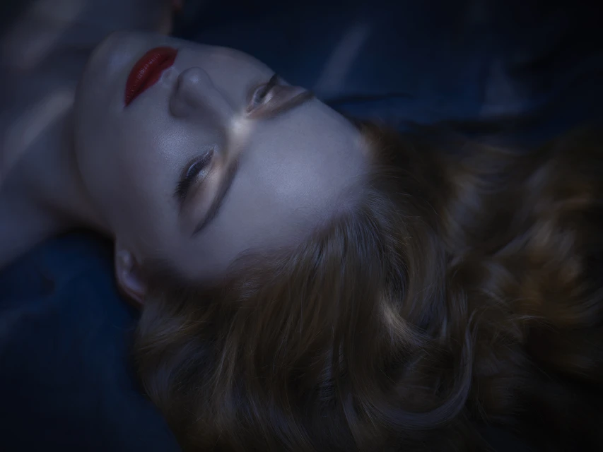 Close-up of a woman with fair skin and red lipstick lying down with reddish-brown wavy hair, illuminated with a blue and white light.