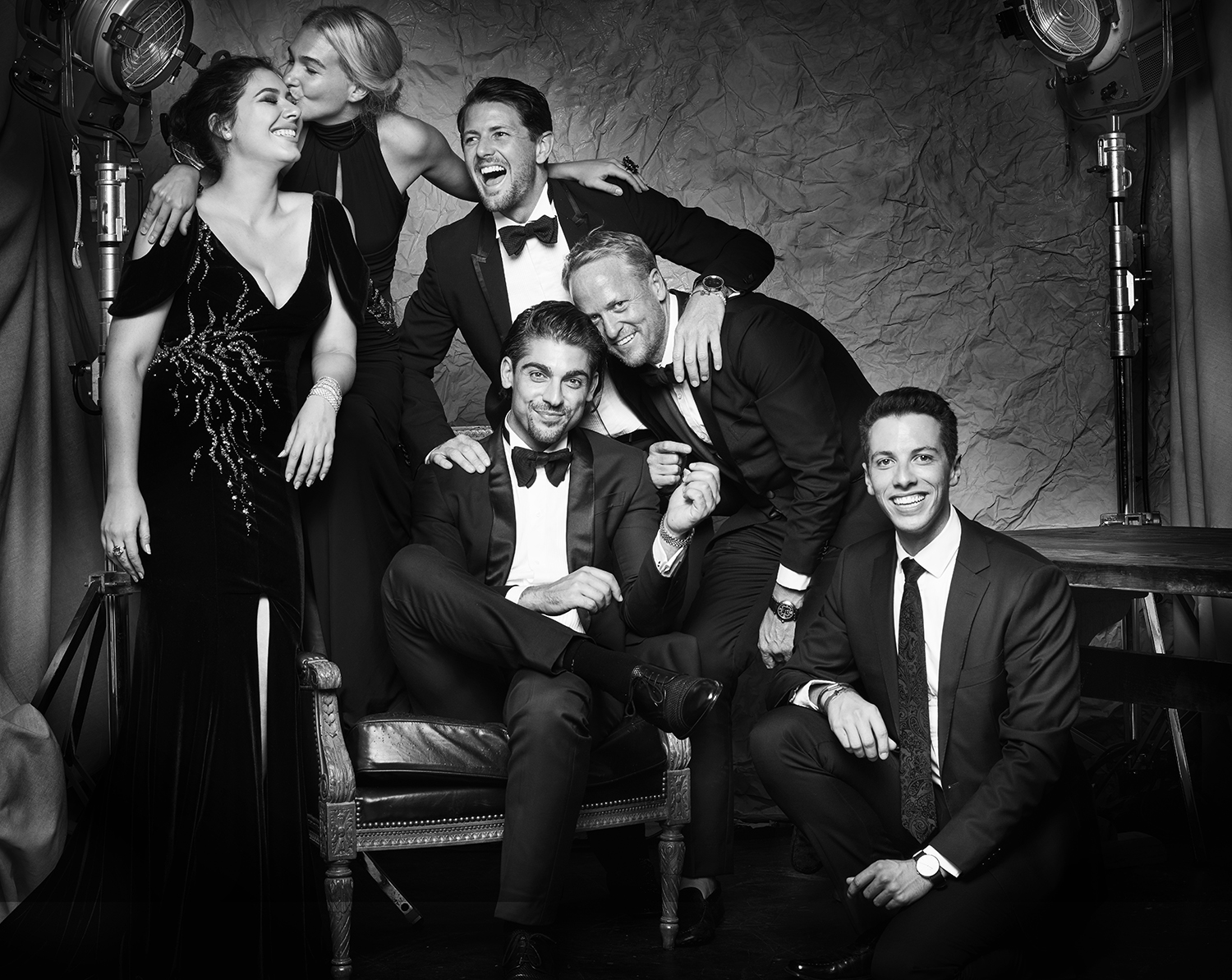 A group of six people pose in formal wear in a black-and-white studio photograph featuring spotlights and an ornate chair.