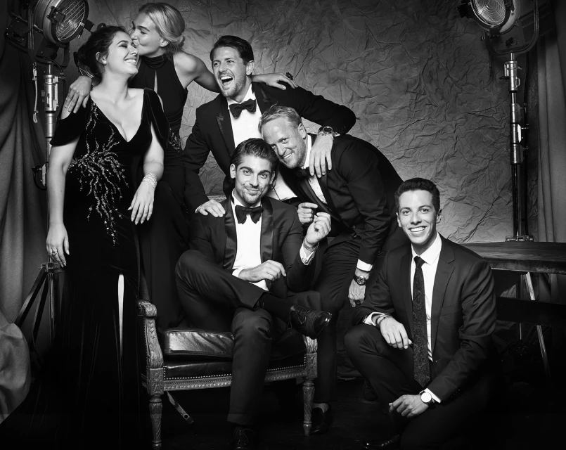 A group of six people pose in formal wear in a black-and-white studio photograph featuring spotlights and an ornate chair.