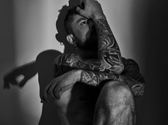 Monochrome portrait of a tattooed man with a beard sitting with his arm raised to his forehead, casting shadows on a plain wall
