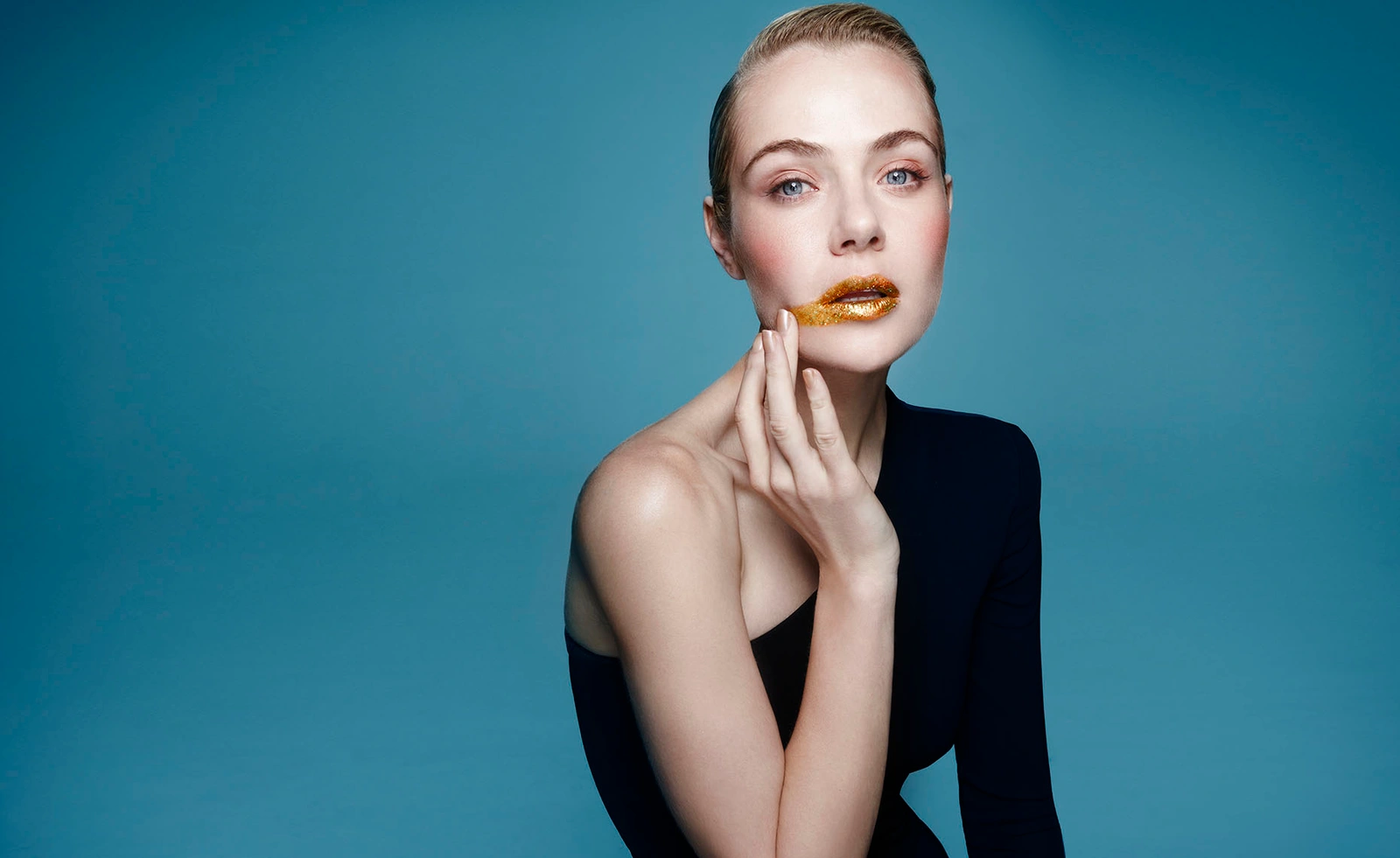 Woman in a black top with gold lip makeup touching her face against a solid blue studio backdrop