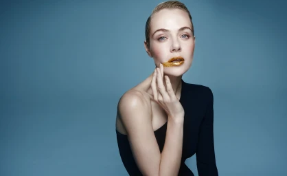 Woman in a black top with gold lip makeup touching her face against a solid blue studio backdrop