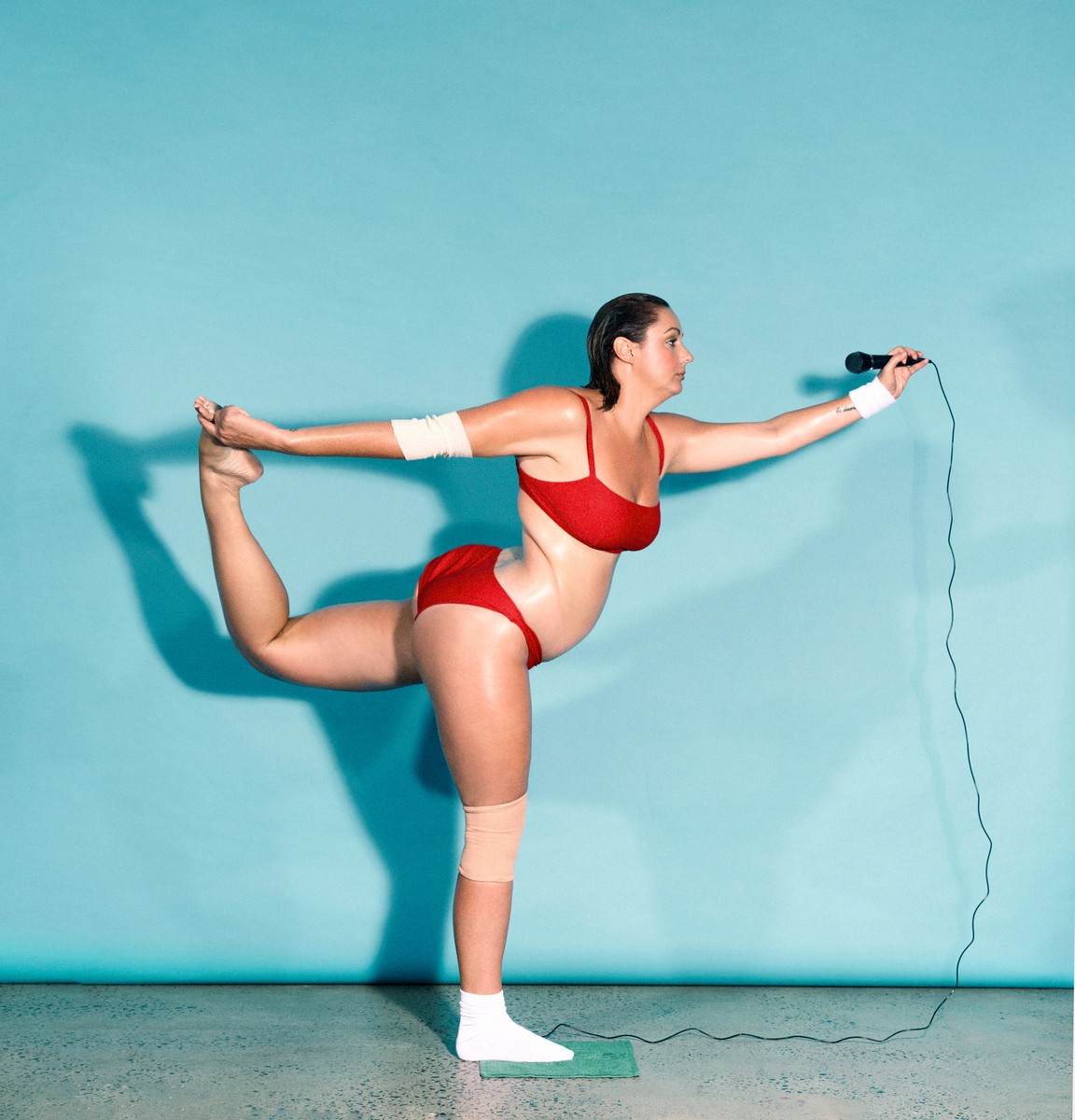 Woman in red swimsuit strikes a yoga pose while holding a microphone with a cord in a minimalist studio setting with a blue backdrop.