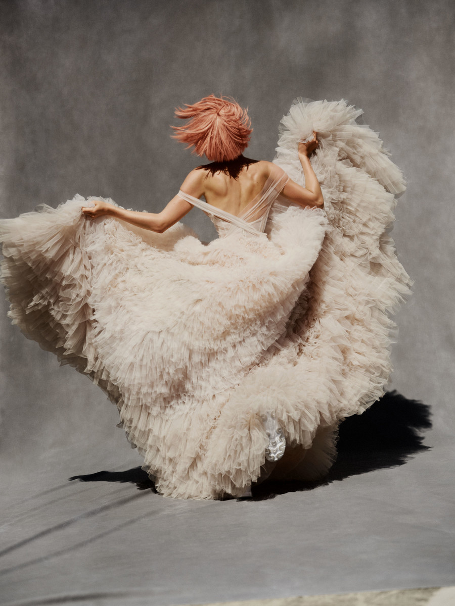 Back view of woman in pale pink tulle gown, hair blowing, against a gray background in a studio setting, fashion forward elegance.