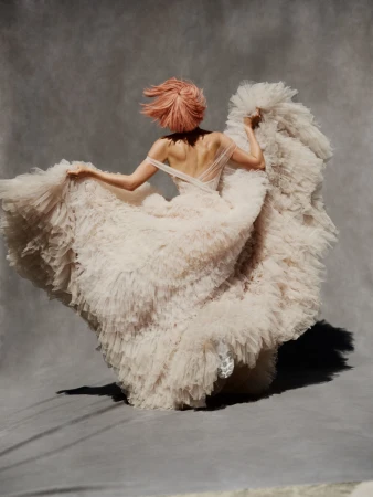 Back view of woman in pale pink tulle gown, hair blowing, against a gray background in a studio setting, fashion forward elegance.