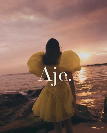 Woman in a yellow puff-sleeve dress standing on a rocky beach at sunset, photographed by Georges Antoni.