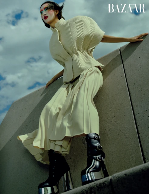 Editorial fashion shot of a woman posing in avant-garde cream-colored outfit and black platform boots with blue sky and clouds in the background.