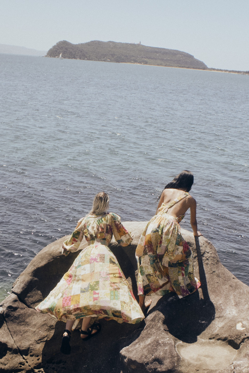 Two women wearing patterned dresses stand on a rock overlooking a sea with a distant island on a sunny day in a fashion shot.