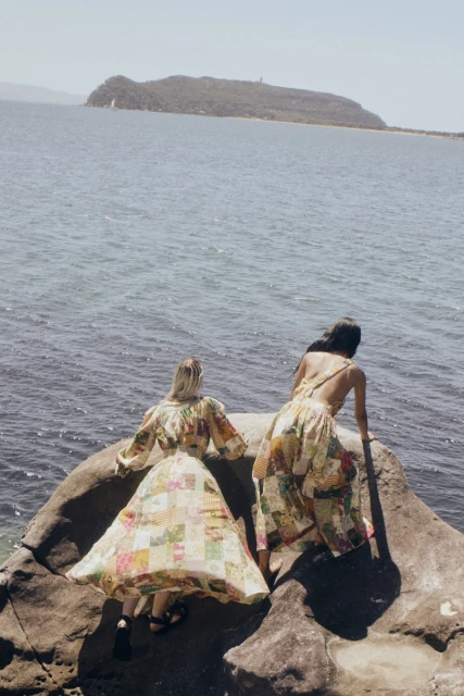Two women wearing patterned dresses stand on a rock overlooking a sea with a distant island on a sunny day in a fashion shot.