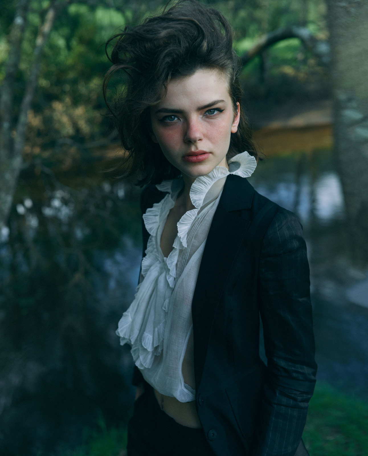 Portrait of a woman with dark hair wearing a black blazer and white ruffled blouse, posing outdoors near a body of water with trees.