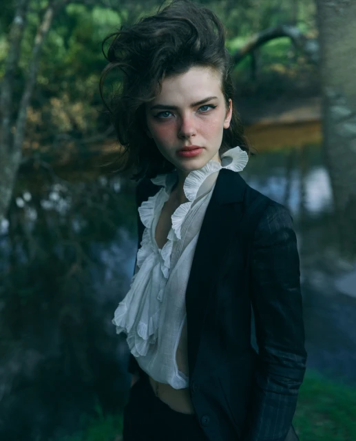 Portrait of a woman with dark hair wearing a black blazer and white ruffled blouse, posing outdoors near a body of water with trees.