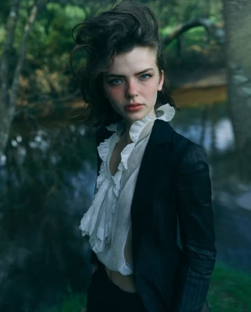 Portrait of a woman with dark hair wearing a black blazer and white ruffled blouse, posing outdoors near a body of water with trees.
