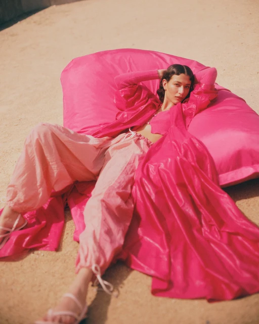 A woman in pink loungewear reclines on a matching pink cushion outdoors, with soft, diffused lighting enhancing the scene's relaxed mood.