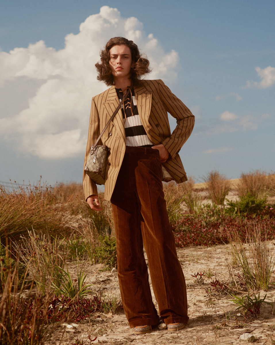 An adult male model with wavy brown hair poses in a field wearing a striped blazer, black and white shirt, and brown corduroy pants.