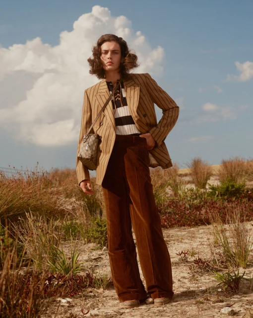 An adult male model with wavy brown hair poses in a field wearing a striped blazer, black and white shirt, and brown corduroy pants.