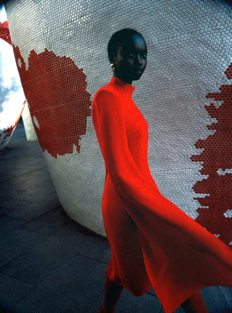Woman in red dress walking beside a patterned wall, showcasing fashion and design, captured in medium shot with natural lighting.