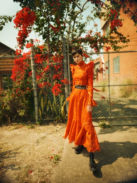 A woman in a flowing orange dress and boots stands on a path near flowering bougainvillea and a fence in a bright outdoor fashion shot.