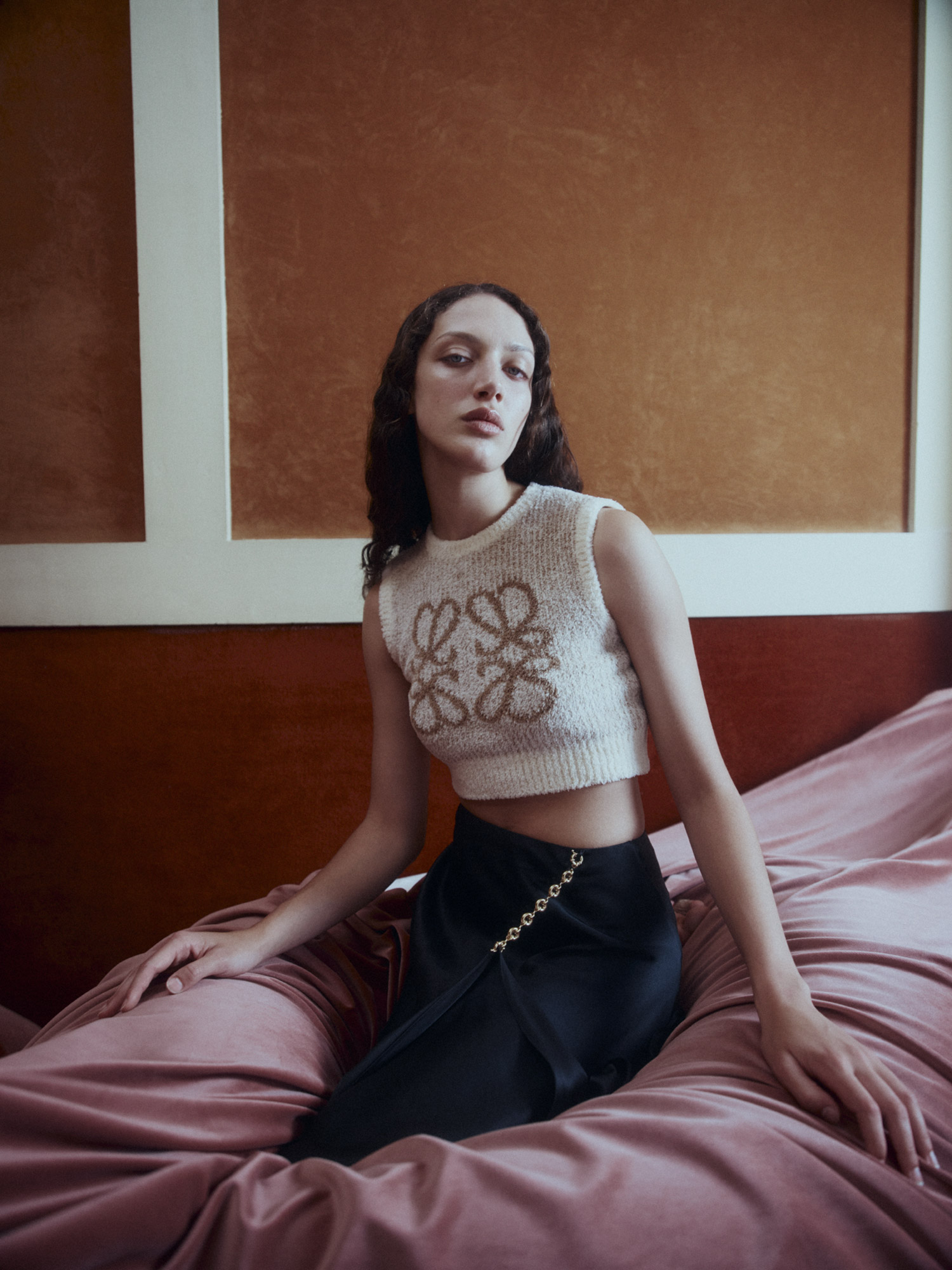 An adult female model with long dark hair sits on a bed wearing a knitted crop top and black skirt in an indoor setting.