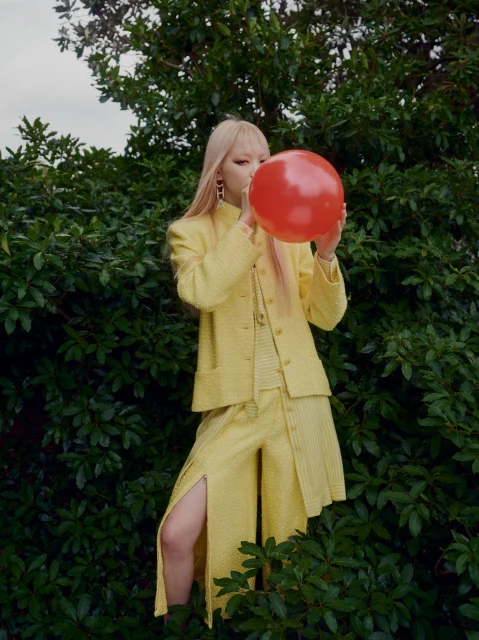Woman with blonde hair blowing up a red balloon, wearing a yellow patterned suit, standing outdoors surrounded by green foliage.