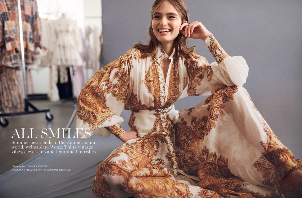 Smiling woman in patterned Zimmermann dress sits in photo studio. Soft, warm light highlights textures of the dress and studio garments rack.
