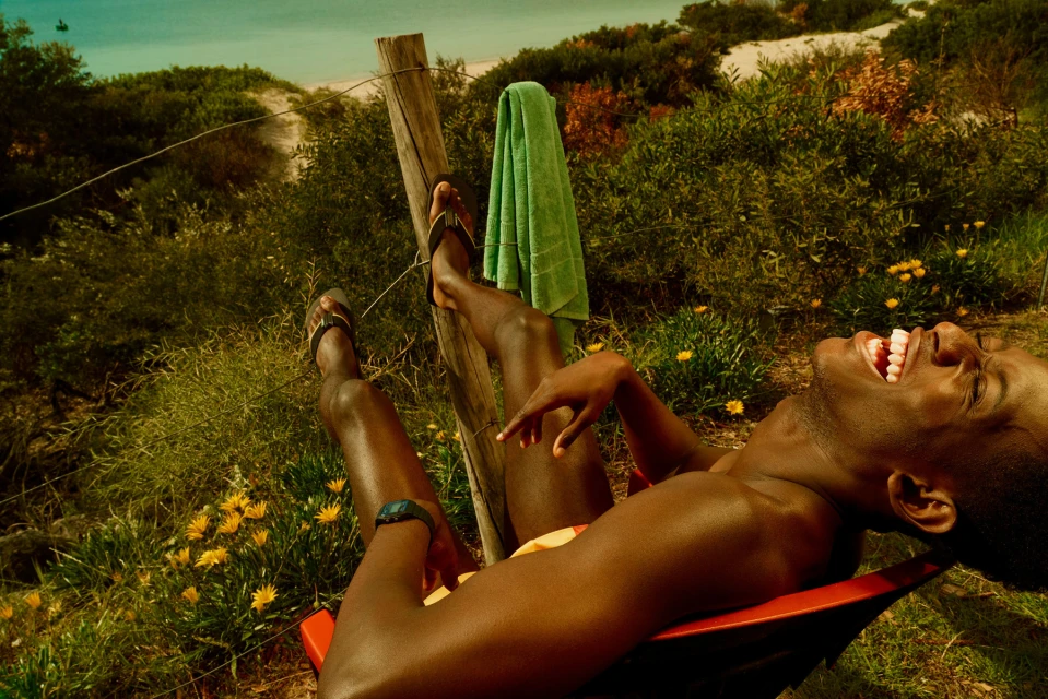 African American man reclining in red chair with towel draped on fence post and vegetation in background, enjoying summer day.