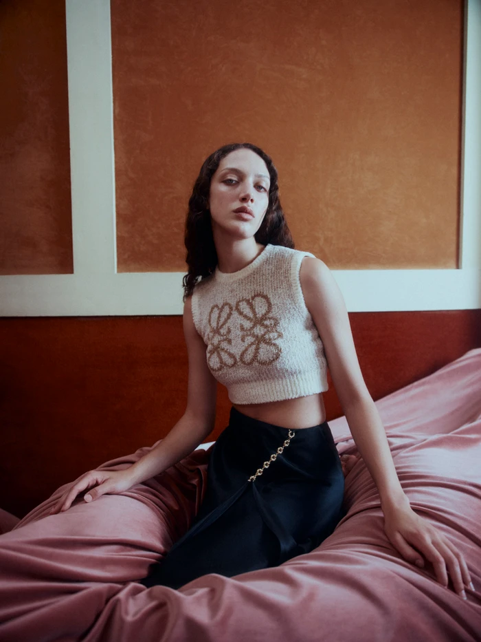 Woman in white knit crop top and black skirt seated on a pink velvet bed by Georges Antoni