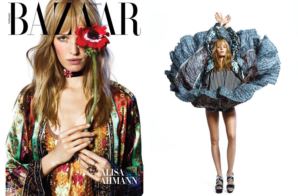 Fashion editorial shot of a blonde model wearing a colorful patterned outfit and holding a red flower, set against a white backdrop.