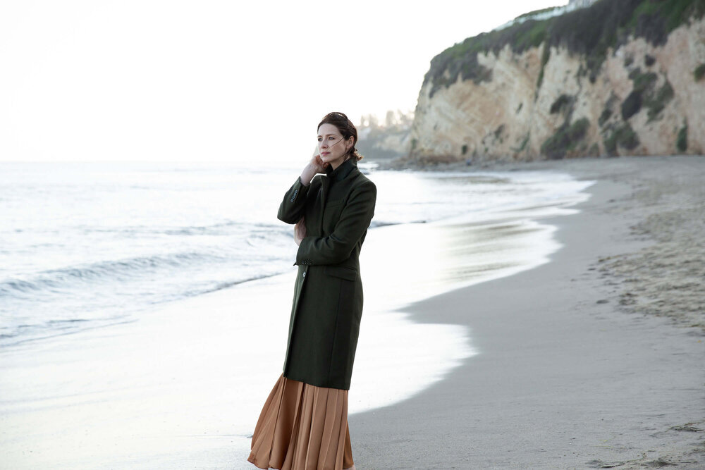Woman in a coat stands on a beach with a tan skirt, ocean, and coastal bluffs in the background on a cloudy day.