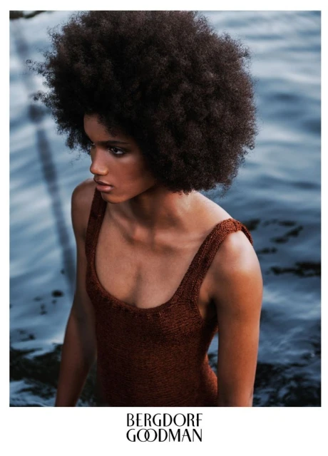 Portrait of a dark-skinned woman with an afro hairstyle wearing a brown swimsuit, standing in a body of water. Fashion editorial.