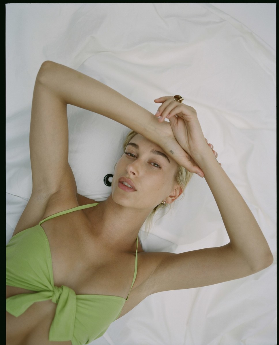 Blonde woman in a light green bikini lies on a white sheet, her arms raised above her head in a bright, indoor studio setting.