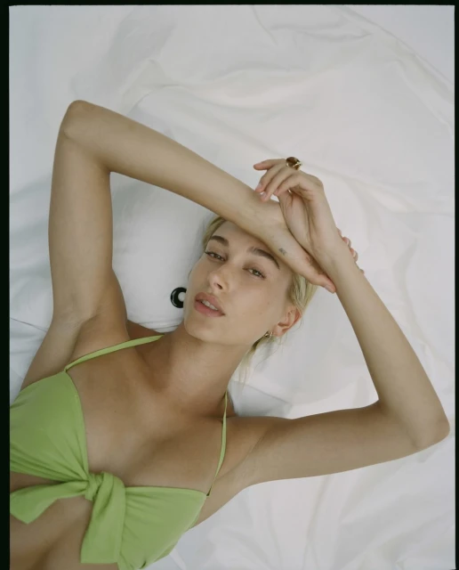 Blonde woman in a light green bikini lies on a white sheet, her arms raised above her head in a bright, indoor studio setting.