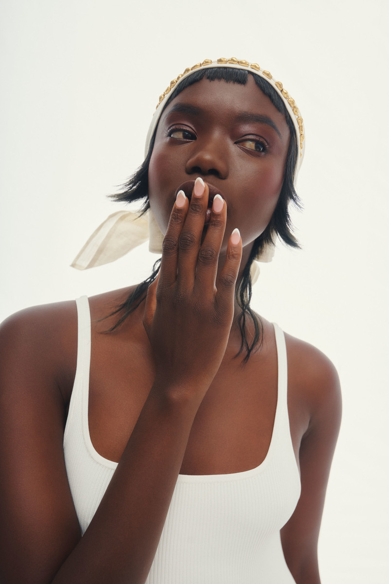 A young Black woman with her hand near her face, wearing a cream-colored headscarf with gold beads and a white ribbed tank top on a white background.