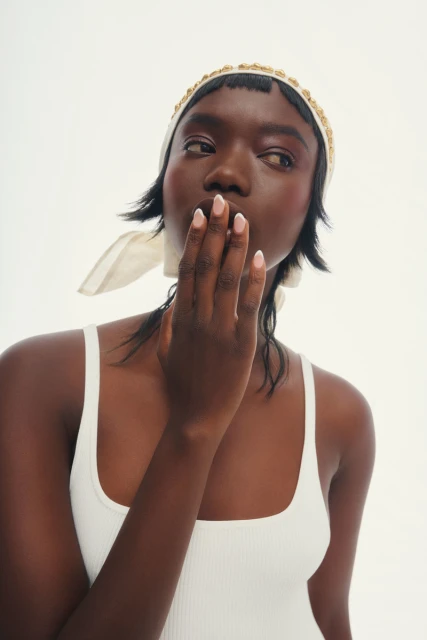 A young Black woman with her hand near her face, wearing a cream-colored headscarf with gold beads and a white ribbed tank top on a white background.