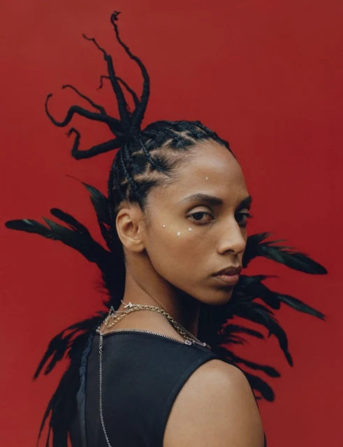 Headshot of an African-American woman against a red background; she is adorned with black feathers, elegant braids, and subtle facial decorations.