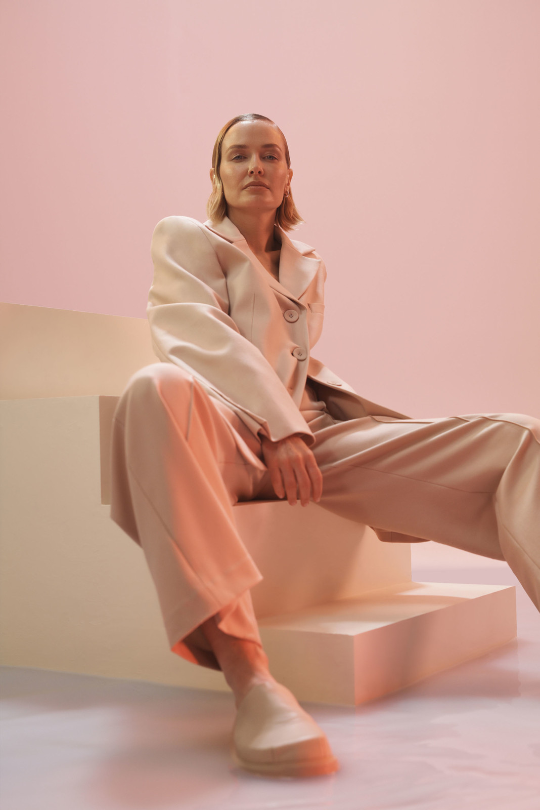 Adult woman in a light beige suit sitting on stairs against a pink background in a studio, gazing directly at the camera.
