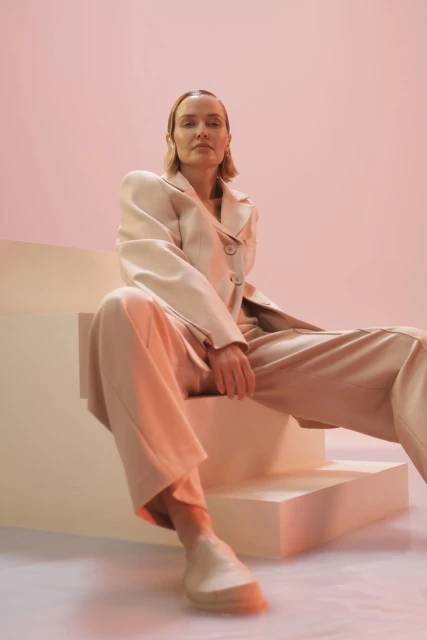 Adult woman in a light beige suit sitting on stairs against a pink background in a studio, gazing directly at the camera.