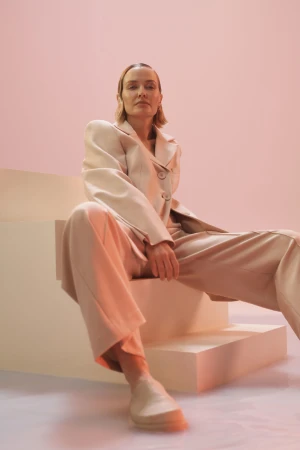 Adult woman in a light beige suit sitting on stairs against a pink background in a studio, gazing directly at the camera.