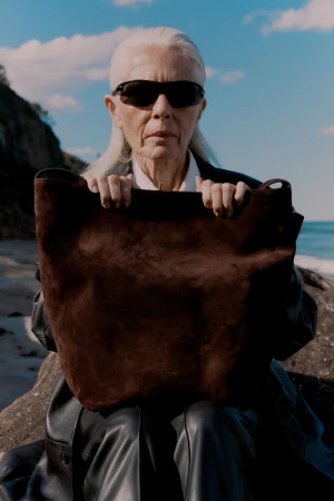 Older woman with white hair wearing sunglasses and a dark coat holding a large brown suede bag by the coast