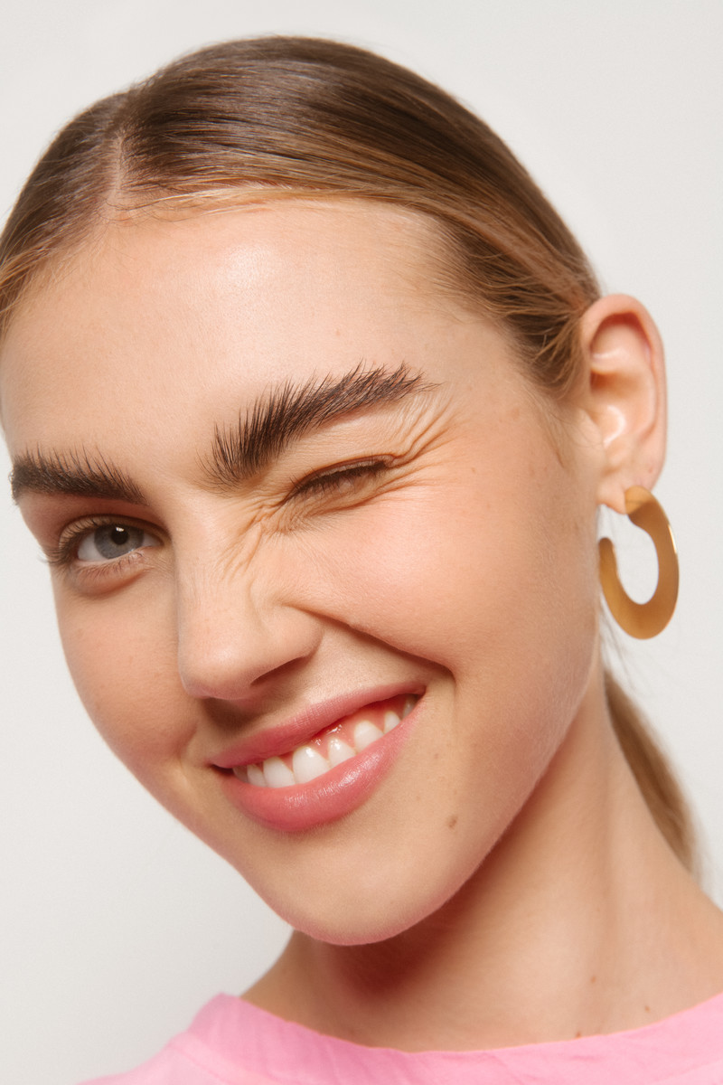 Close-up portrait of a young adult woman with brown hair pulled back, hoop earring, wearing pink top, winking with a smile against white.