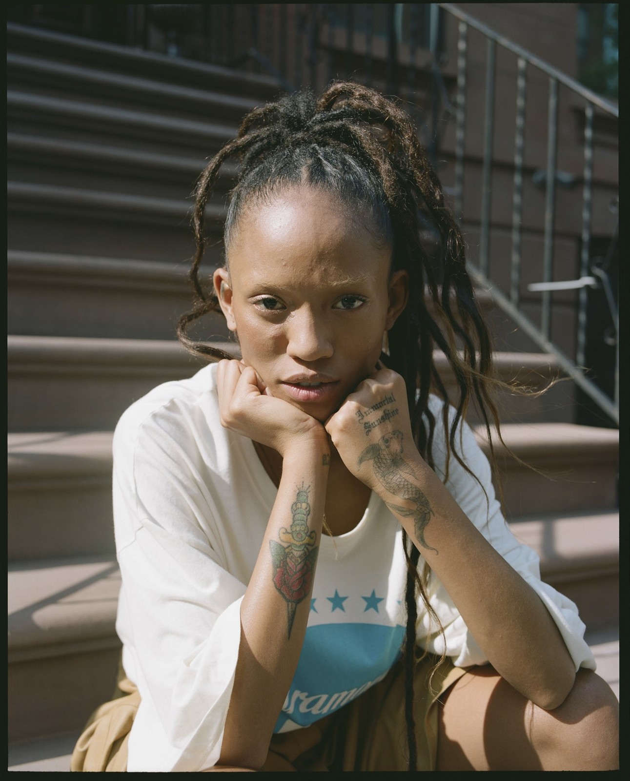 Adult female seated on brownstone steps with a neutral expression, wearing a white t-shirt with blue graphic and khaki shorts.