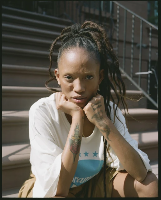 Adult female seated on brownstone steps with a neutral expression, wearing a white t-shirt with blue graphic and khaki shorts.