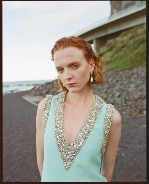Elegant red-haired woman in an embellished dress standing on an asphalt path, with a bridge and the ocean in the background.