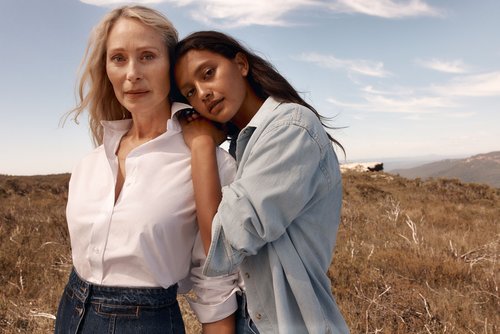 Outdoor fashion shot of a mature woman and a young woman in denim and white shirts, posing closely in a grassy field on a sunny day.