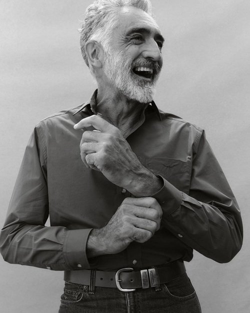 Monochrome portrait of a laughing, bearded, mature adult male in a collared shirt and jeans, adjusting his cuff, with a neutral backdrop.
